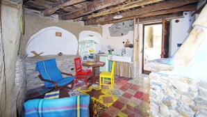 Une pièce rustique aux murs de pierre, au sol carrelé et aux meubles en bois. On y trouve une petite table avec des chaises, un coin cuisine et une porte-fenêtre donnant sur l’extérieur.