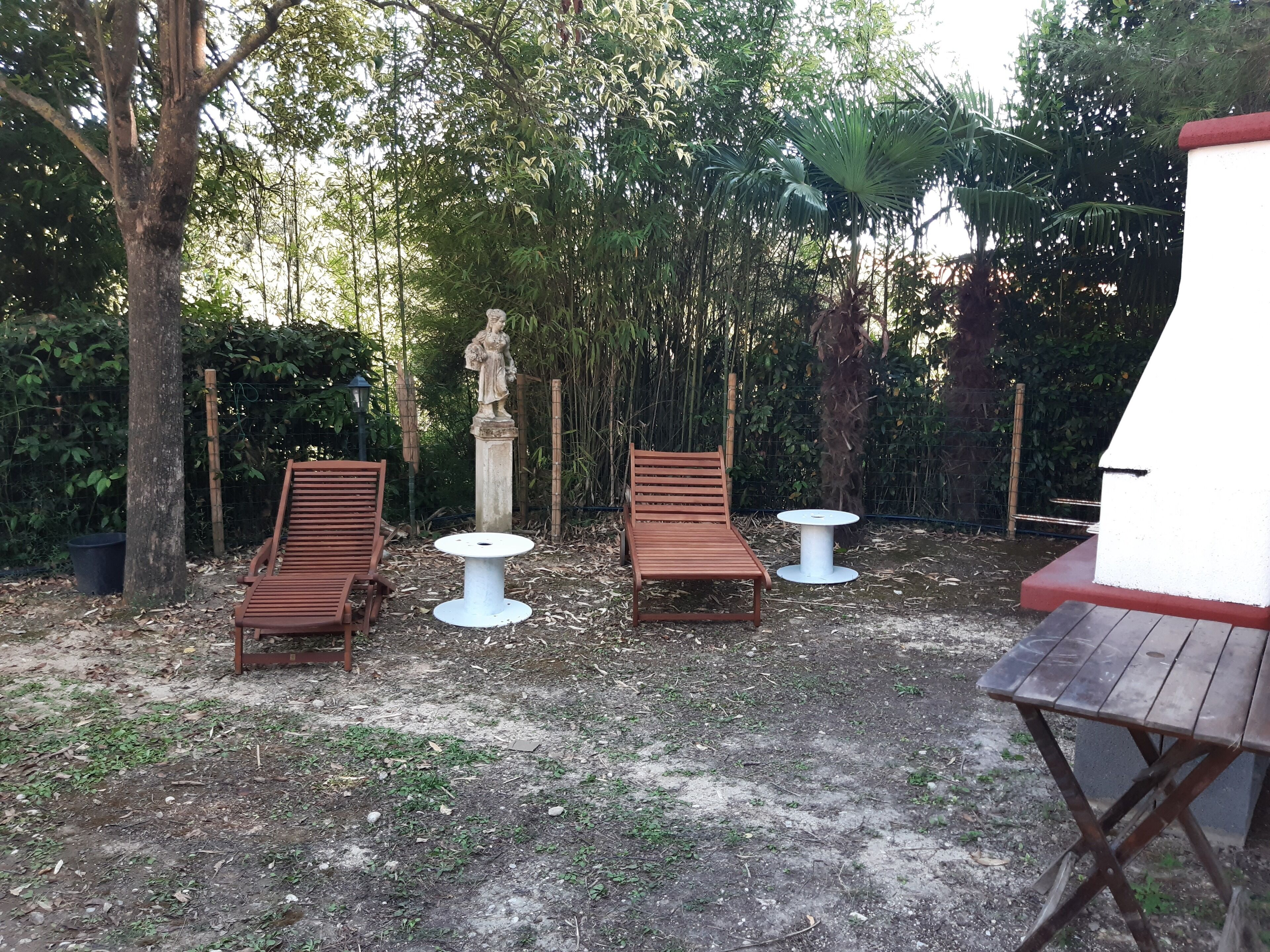 Ein Sitzbereich im Freien mit zwei Holzliegen und zwei kleinen runden Tischen, umgeben von Bäumen und einer Statue.