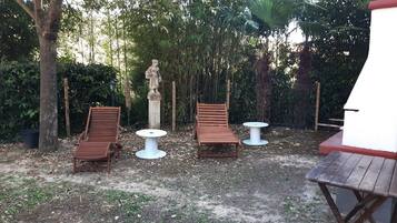 Ein Sitzbereich im Freien mit zwei Holzliegen und zwei kleinen runden Tischen, umgeben von Bäumen und einer Statue.