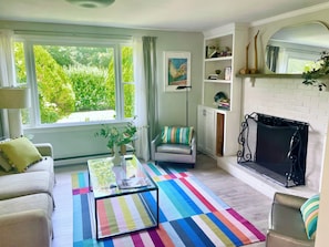 A living room with a sofa, armchair, and a colorful striped rug. There is a fireplace and a bookshelf with books and decorative items.