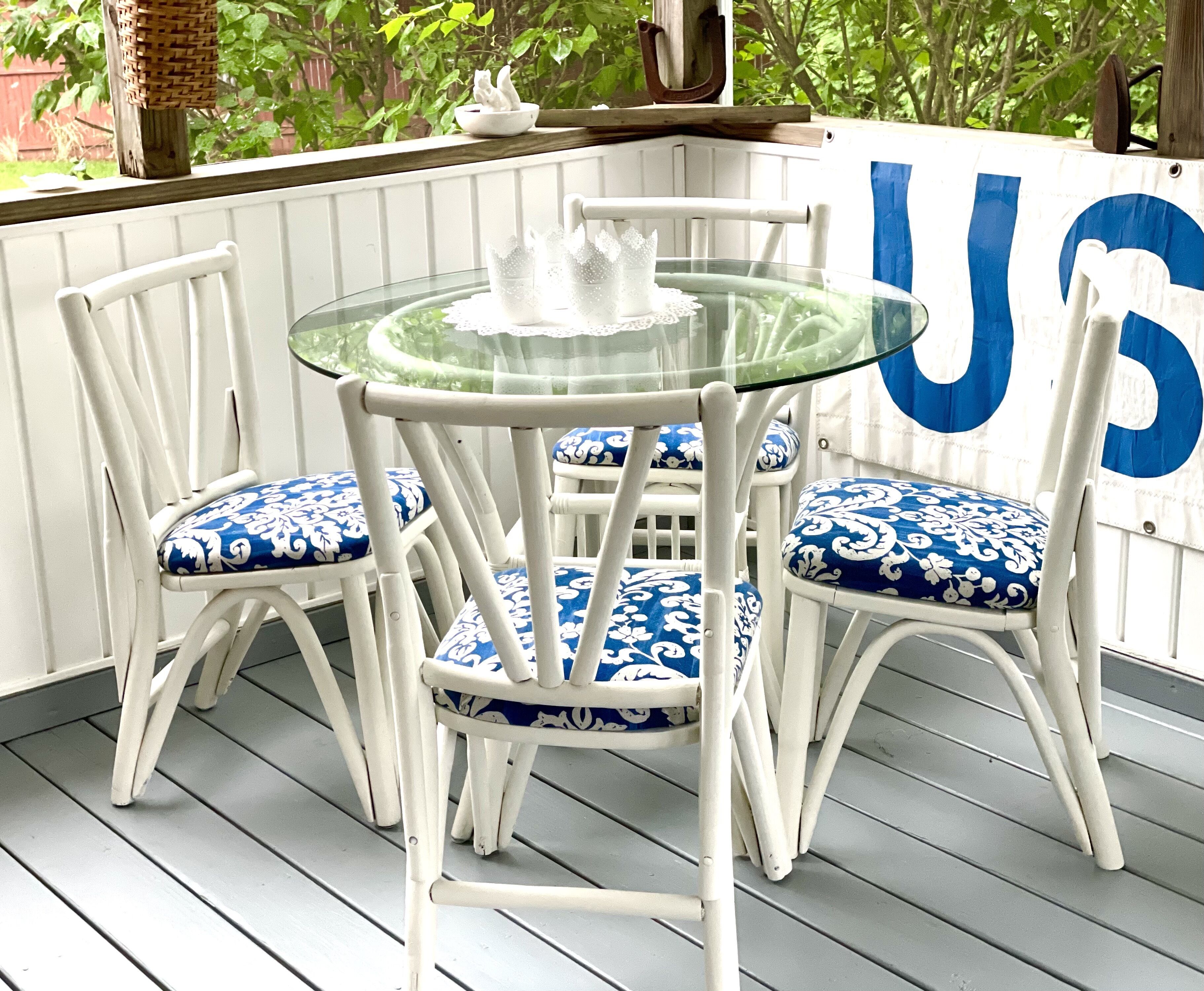 Un área de estar exterior con muebles blancos, una mesa y sillas de vidrio, un comedero para pájaros de mimbre y una cerca blanca con una gran letra 'U' azul.