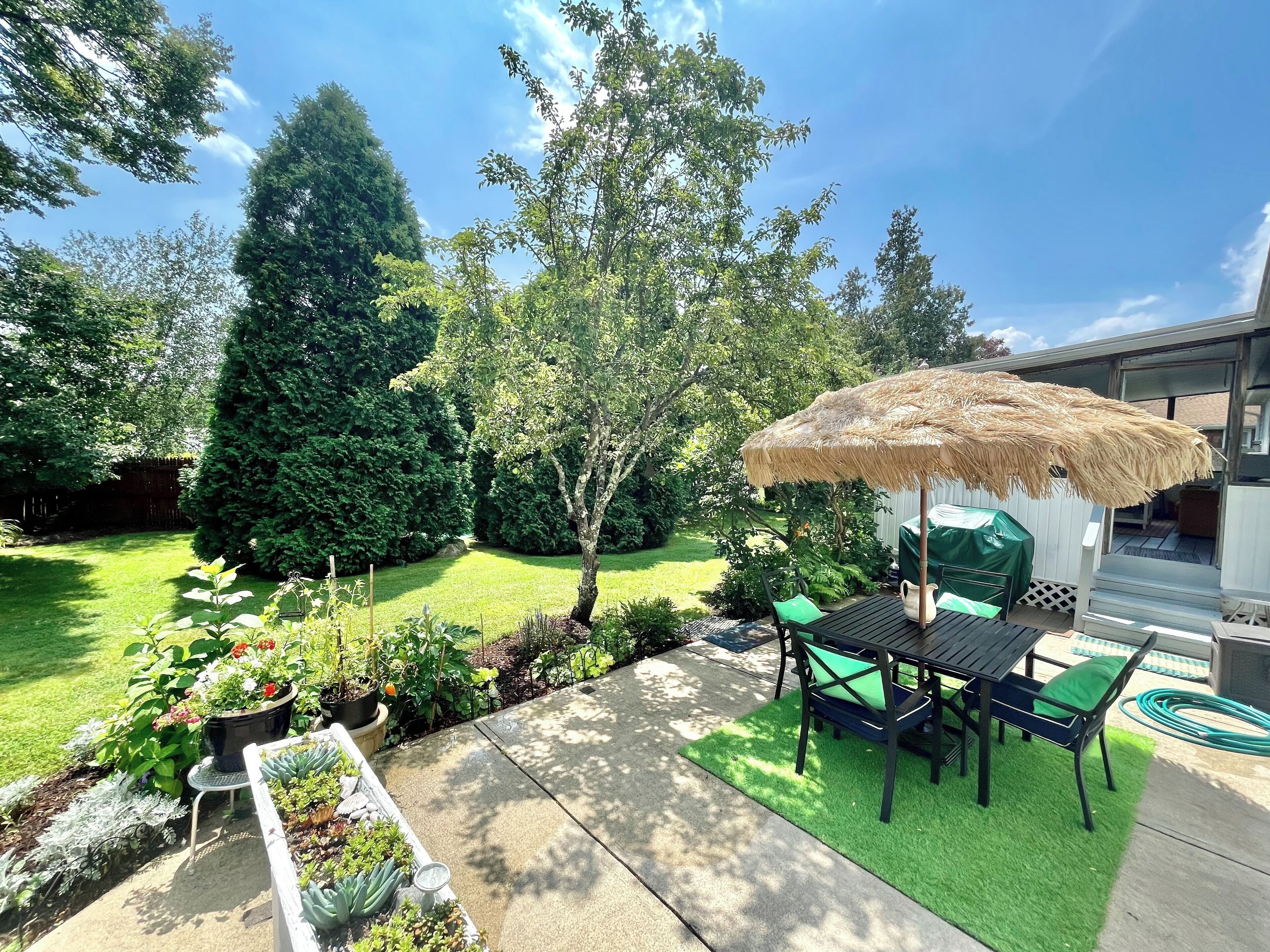 Un patio con mesa y sillas, un parasol de paja y un jardín con diversas plantas y césped.