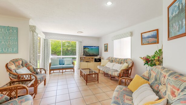 A cozy living room with wicker furniture, a television, and a large window offering a view of greenery. - 22 Bussey Street Bargara (BARGARA)
