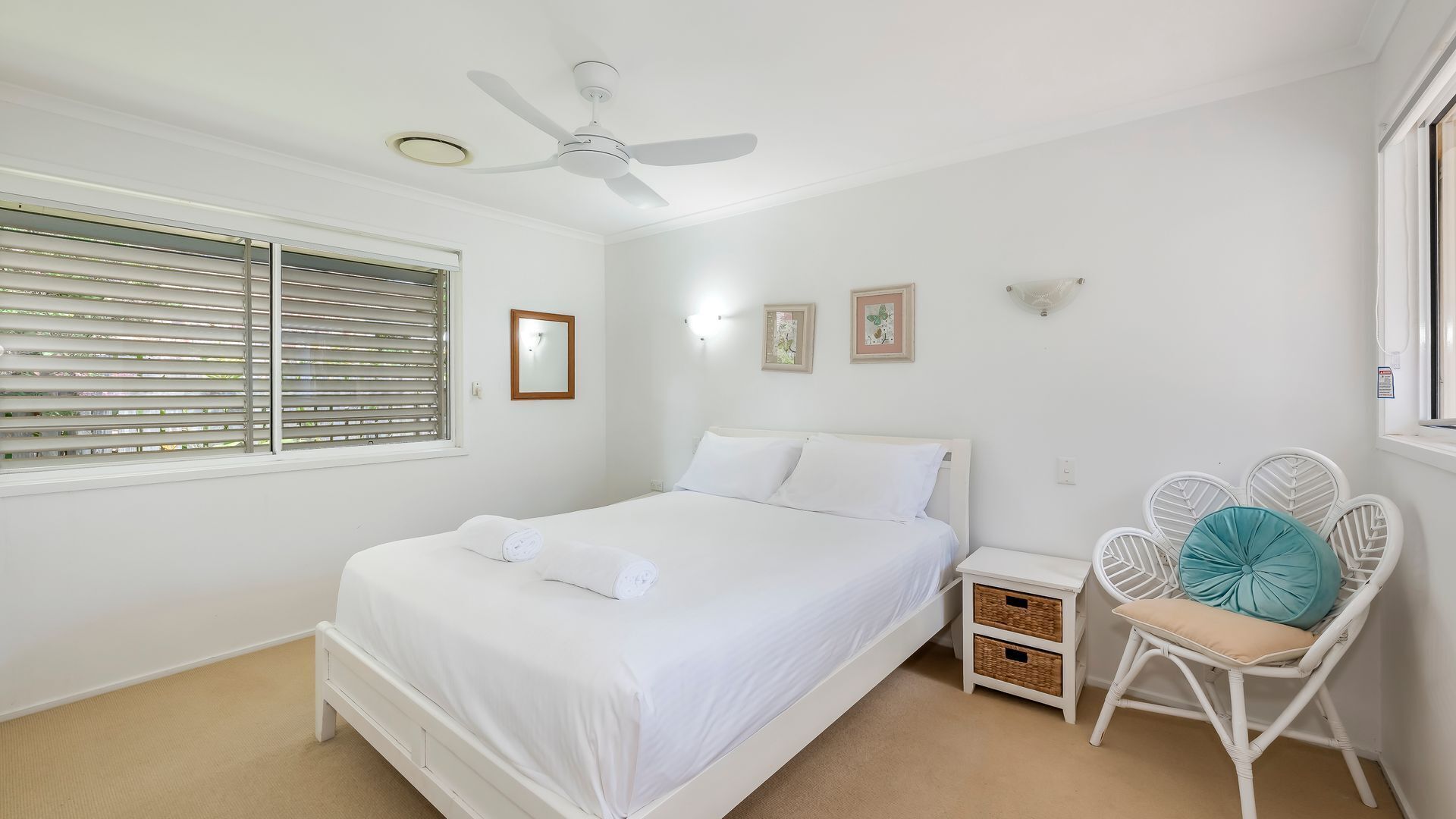 A bedroom with a bed, a ceiling fan, a window with blinds, a nightstand, a chair, and wall-mounted lights.