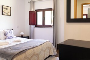 Une chambre à coucher avec un lit, une table de chevet, une lampe, une fenêtre avec des rideaux, un miroir et une citation encadrée accrochée au mur.