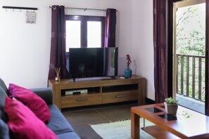 A living room with a TV on a wooden stand, a sofa with cushions, a small table with a plant, and a balcony with a railing.