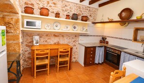 Uma cozinha com mesa de madeira, cadeiras, micro-ondas e diversos pratos de cerâmica nas prateleiras.