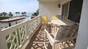 A balcony with a view of the beach and pool, white furniture, and a patterned floor.