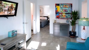 Ein Wohnzimmer mit Fernseher, einem blauen Sessel, einer Holzkommode und einem Bett im Hintergrund.