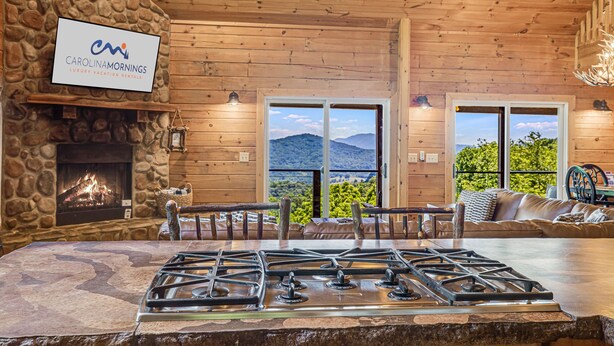 Ein gemütliches Blockhaus mit Kamin aus Stein, einer Steinwand, einer Holzbalkendecke und Bergblick durch große Fenster.