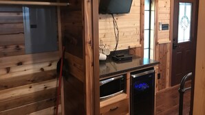 Interior de cabana de madeira com televisão na parede, uma pequena mesa com controle remoto e geladeira embutida.