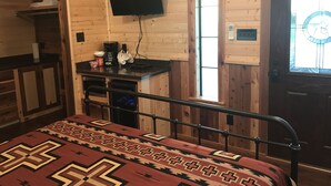 Um quarto de cabana acolhedor com uma cama, uma escrivaninha com um computador, uma TV fixada na parede e uma porta de madeira com um janelão.