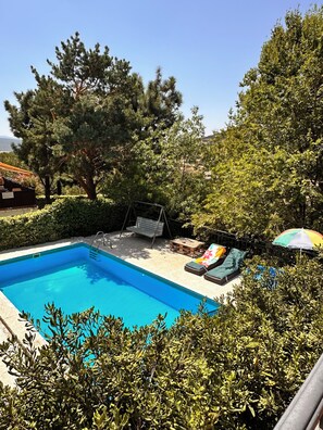 A swimming pool with a swing chair and lounge chairs, surrounded by greenery and trees.