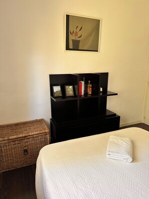 A hotel room with a bed, a bedside table, a small shelf with items, a wicker basket, and a painting on the wall.