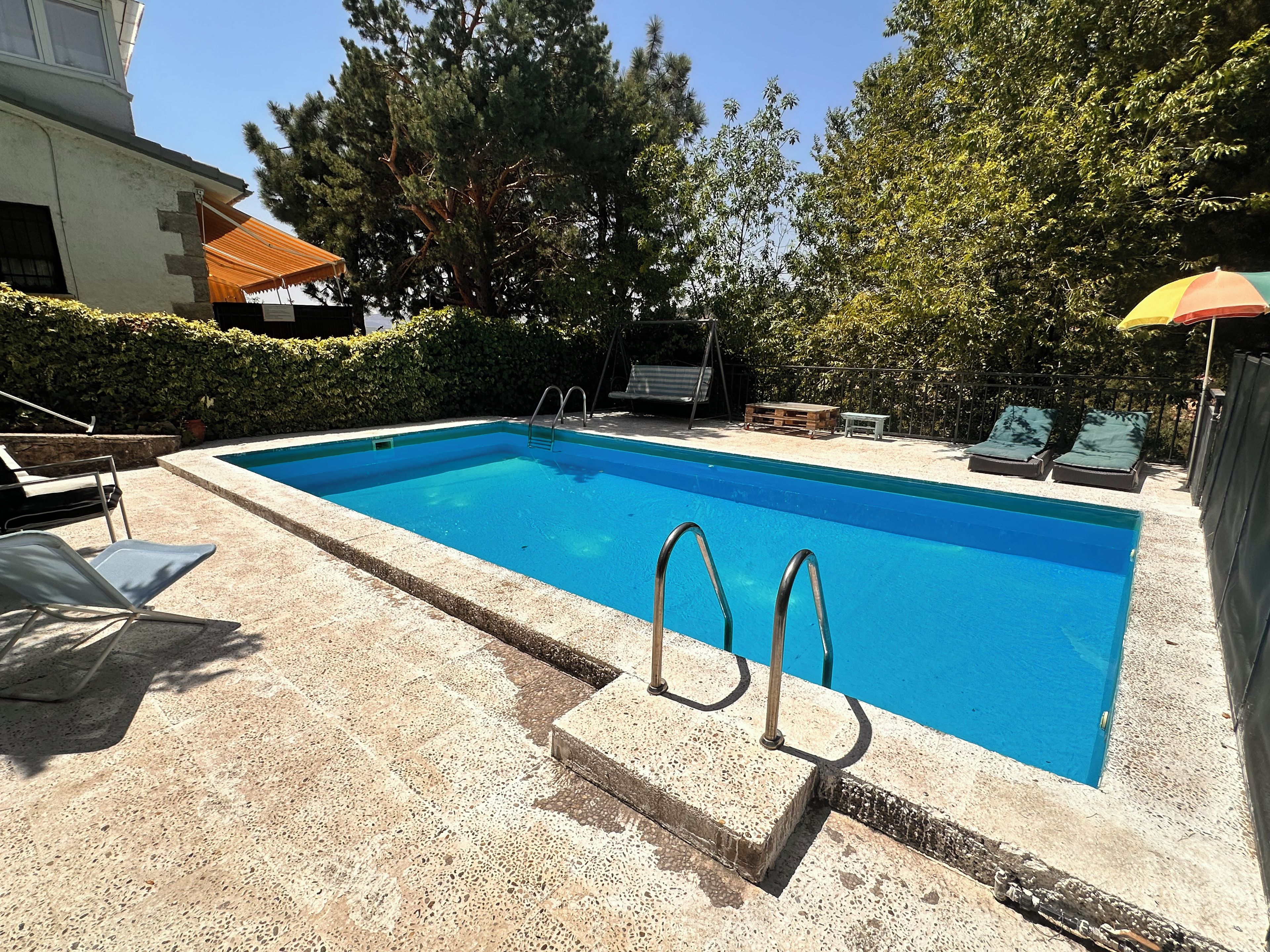 Ein rechteckiger Swimmingpool mit einer Leiter, umgeben von einer Betonplattform. Liegestühle und ein kleiner Tisch mit einer Holzbank befinden sich in der Nähe.