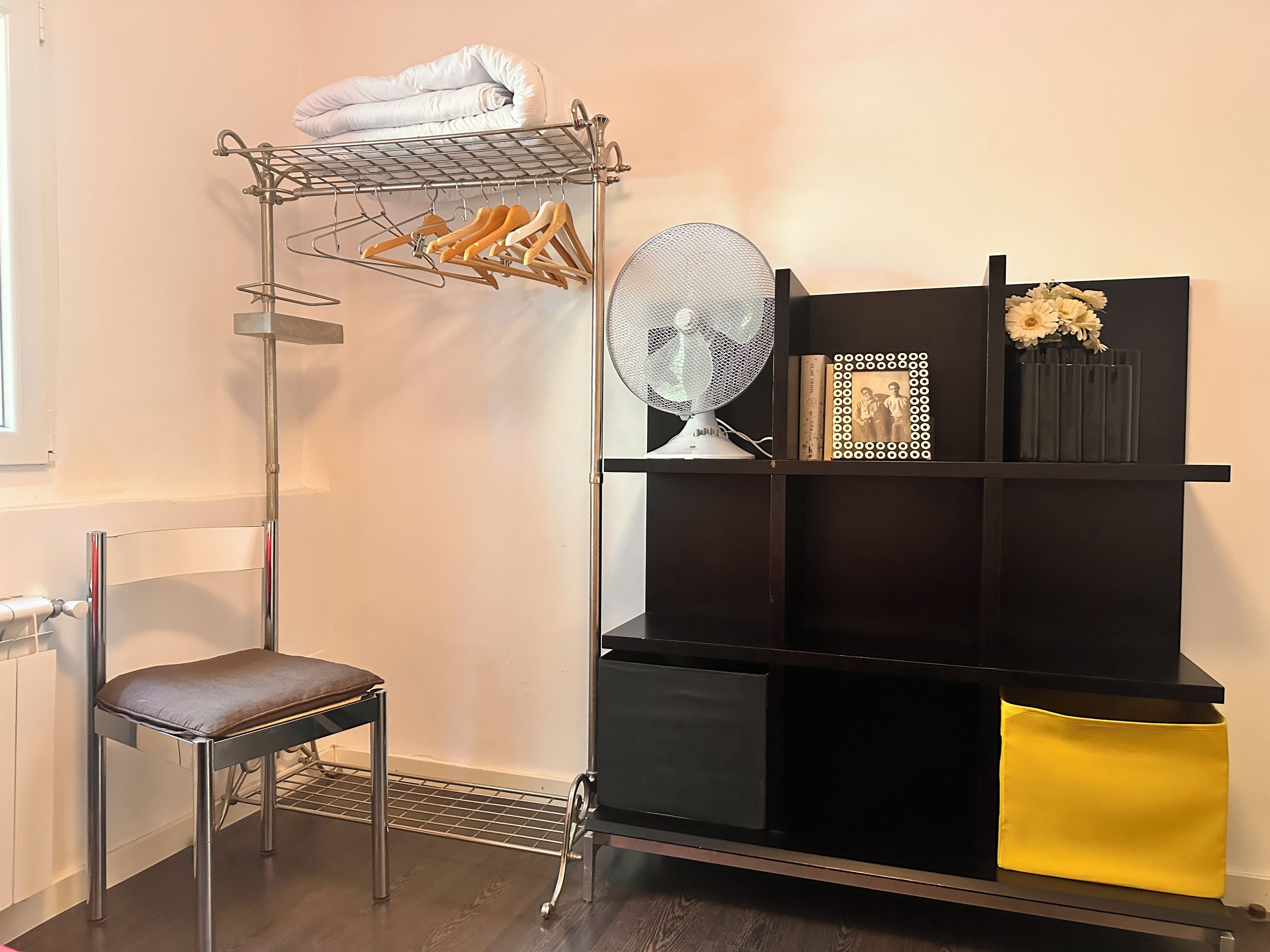 Une pièce comprenant un présentoir métallique où sont pliées des serviettes, une chaise, un ventilateur et une étagère noire ornée d’objets décoratifs.