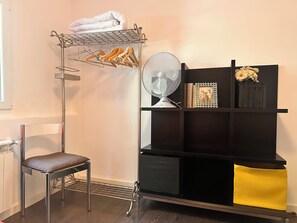 Una habitación con un perchero metálico que contiene toallas dobladas, una silla, un ventilador y un estante negro con objetos decorativos.