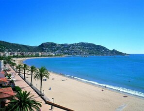 A coastal beach with a clear view of the sea, a sandy shore, and a hillside in the background.