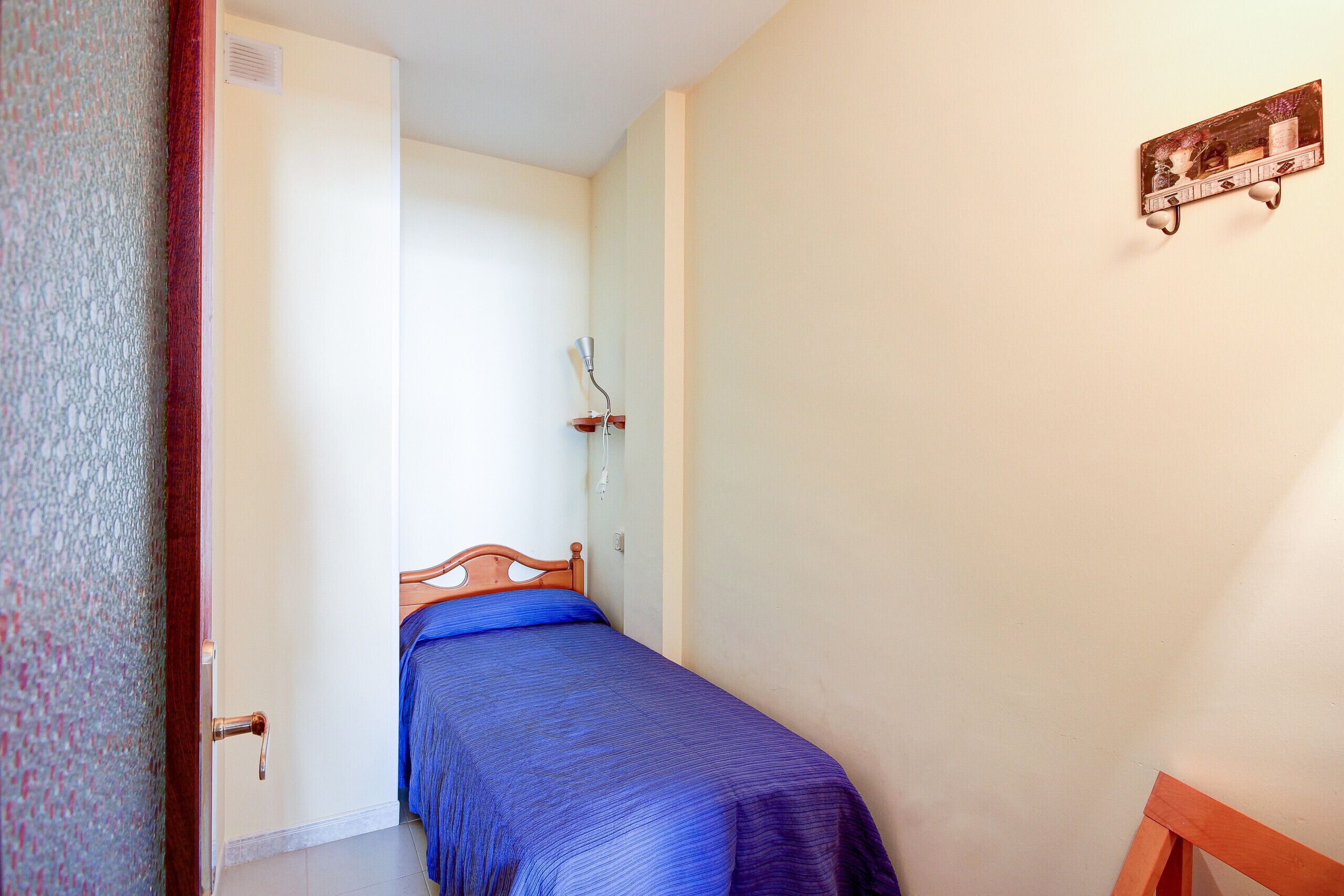 A small, single-bed room with a wooden headboard, a bedside table, and a wall-mounted shelf.