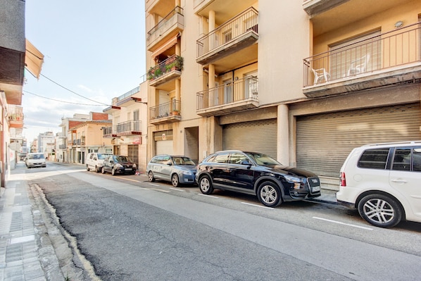 A street with parked cars, a building with balconies, and a closed shutter.