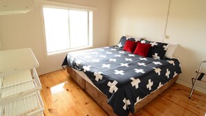 Une chambre avec un plancher en bois, un lit recouvert d’une couvre-lit à motifs croisés noirs et blancs, des oreillers rouges et une table de chevet blanche.