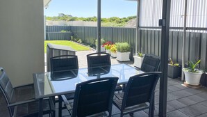 A covered patio area with a glass table and chairs, potted plants, and a view of a fenced yard.