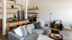 A modern living room with a grey sofa, a coffee table, and a kitchen area with wooden shelves.