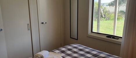 Un dormitorio con colcha a cuadros, una toalla enrollada y una ventana con vistas a un jardín.