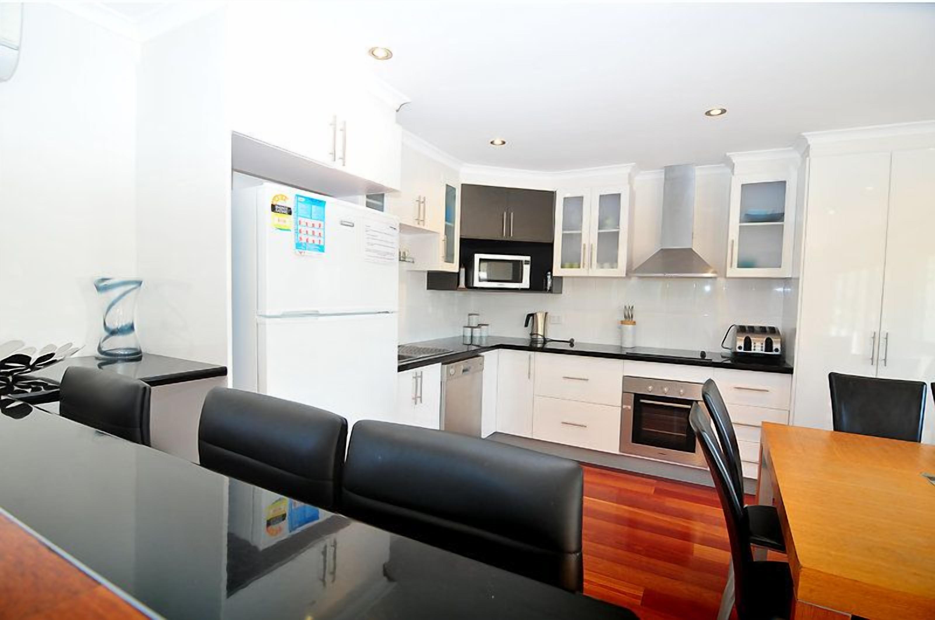 A modern kitchen with white cabinets, a microwave, and a stove. There is a dining area with a wooden table and black chairs.