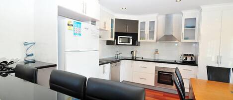 A modern kitchen with white cabinets, a microwave, and a stove. There is a dining area with a wooden table and black chairs.