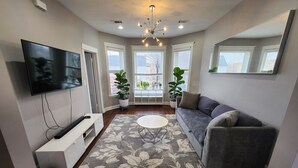 Ein Wohnzimmer mit Flachbildfernseher, grauem Sofa, floralem Teppich und einer Deckenleuchte.