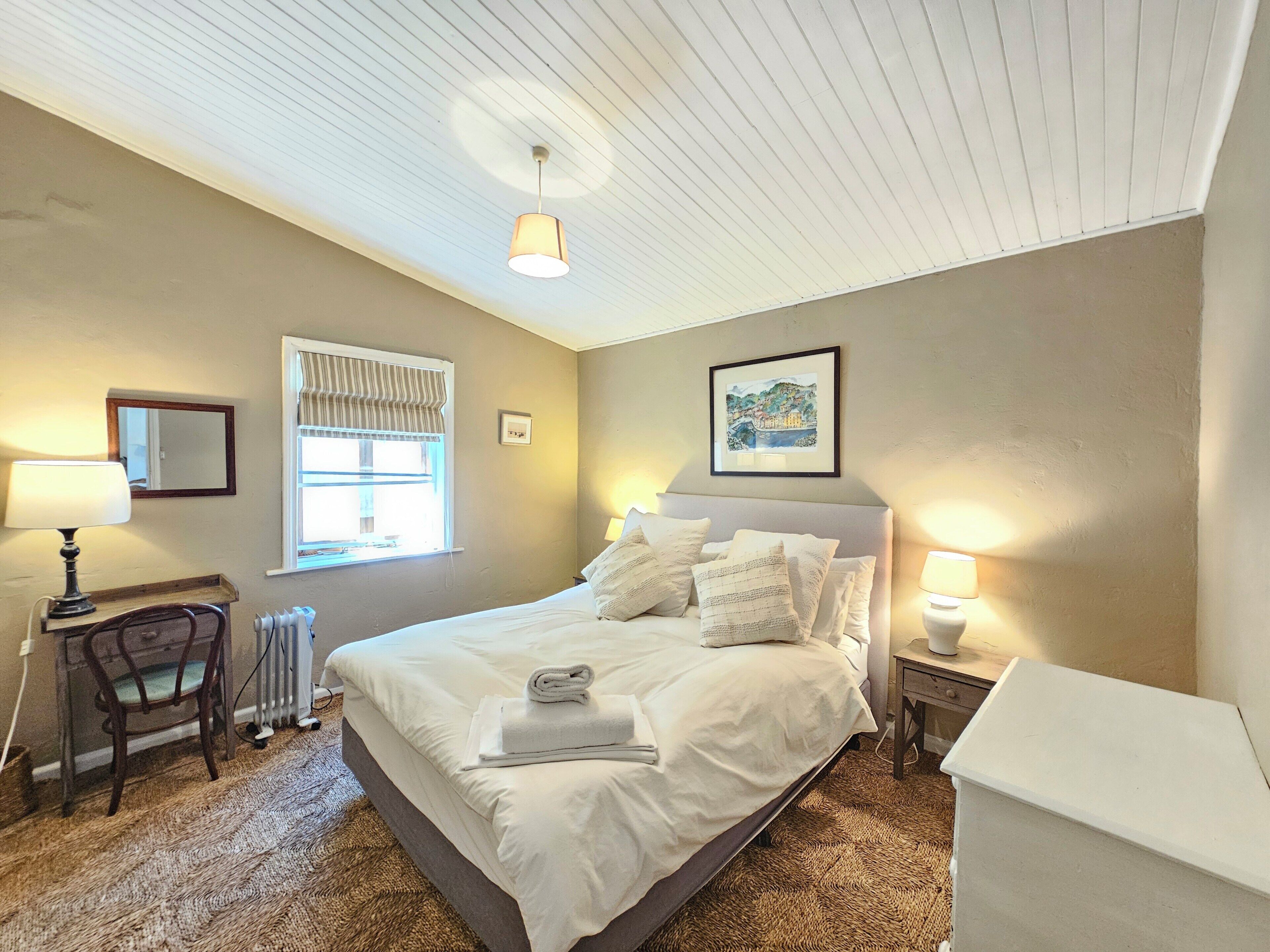 A bedroom with a bed, two bedside lamps, a radiator, a chair, a window with curtains, a framed picture on the wall, and a ceiling light.