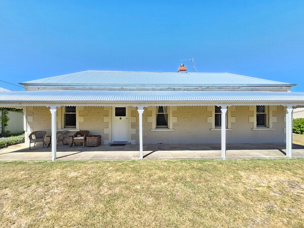 A two-story building with a corrugated metal roof, a covered porch with chairs, and a small window. - Stone Cottage - Gorgeous cottage charm, in the heart of Robe Town (Robe)