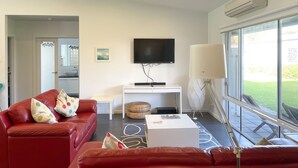 A modern living room with a red sofa, a white coffee table, and a flat-screen TV mounted on the wall. There is a large window providing a view of the outdoors. - Sun on Sanders - Modern 2 br house close to main street. Pet Friendly Outside (Robe)