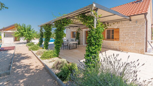 Een stenen huis met een overdekt terras, zitplaatsen buiten en weelderige begroeiing.