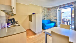 A compact kitchen with a sink, stove, and a small refrigerator. There is a blue sofa and a dining table with chairs. The room has a stone wall and a view of mountains through a glass door. - LUCIA by KlabHouse-Central  lakeview w/AC  (Bellagio)