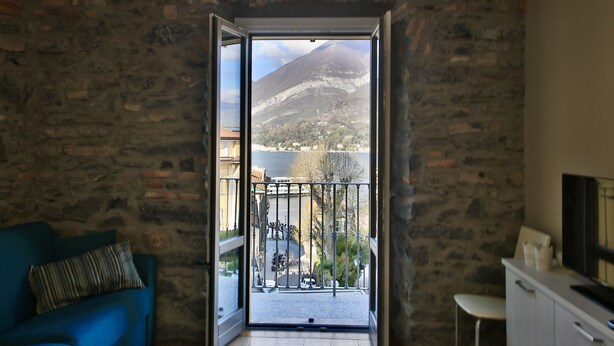 A room with a stone wall, a blue sofa, a white cabinet, and a balcony with a view of a mountain and a lake. - LUCIA by KlabHouse-Central  lakeview w/AC  (Bellagio)