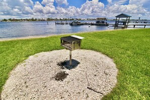 Une propriété située au bord de l’eau, dotée d’un quai, d’une petite structure abritée et d’une zone herbeuse avec un chemin de gravier menant à un espace barbecue en métal.