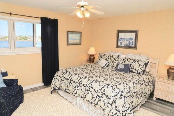 A bedroom with a bed, a nightstand, a lamp, a ceiling fan, a window with a view, and a curtain.