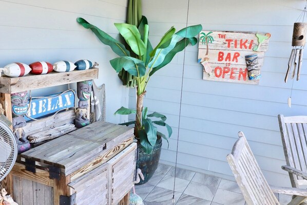 Et skilt med tiki-tema, træflipper og en potteplante på en veranda.