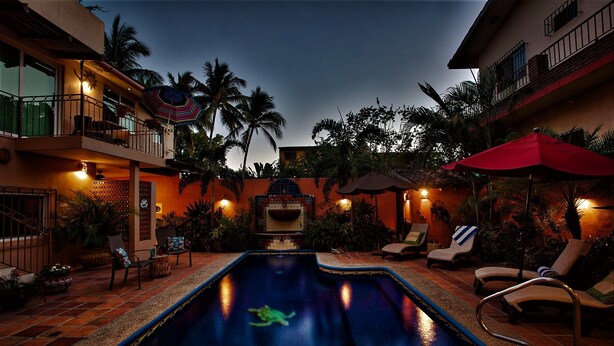 Una piscina con sillones de descanso y sombrillas, rodeada de palmeras y un edificio con balcones.