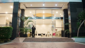 Entrada de hotel con fachada de vidrio, una recepción y una escalera que sube al vestíbulo.