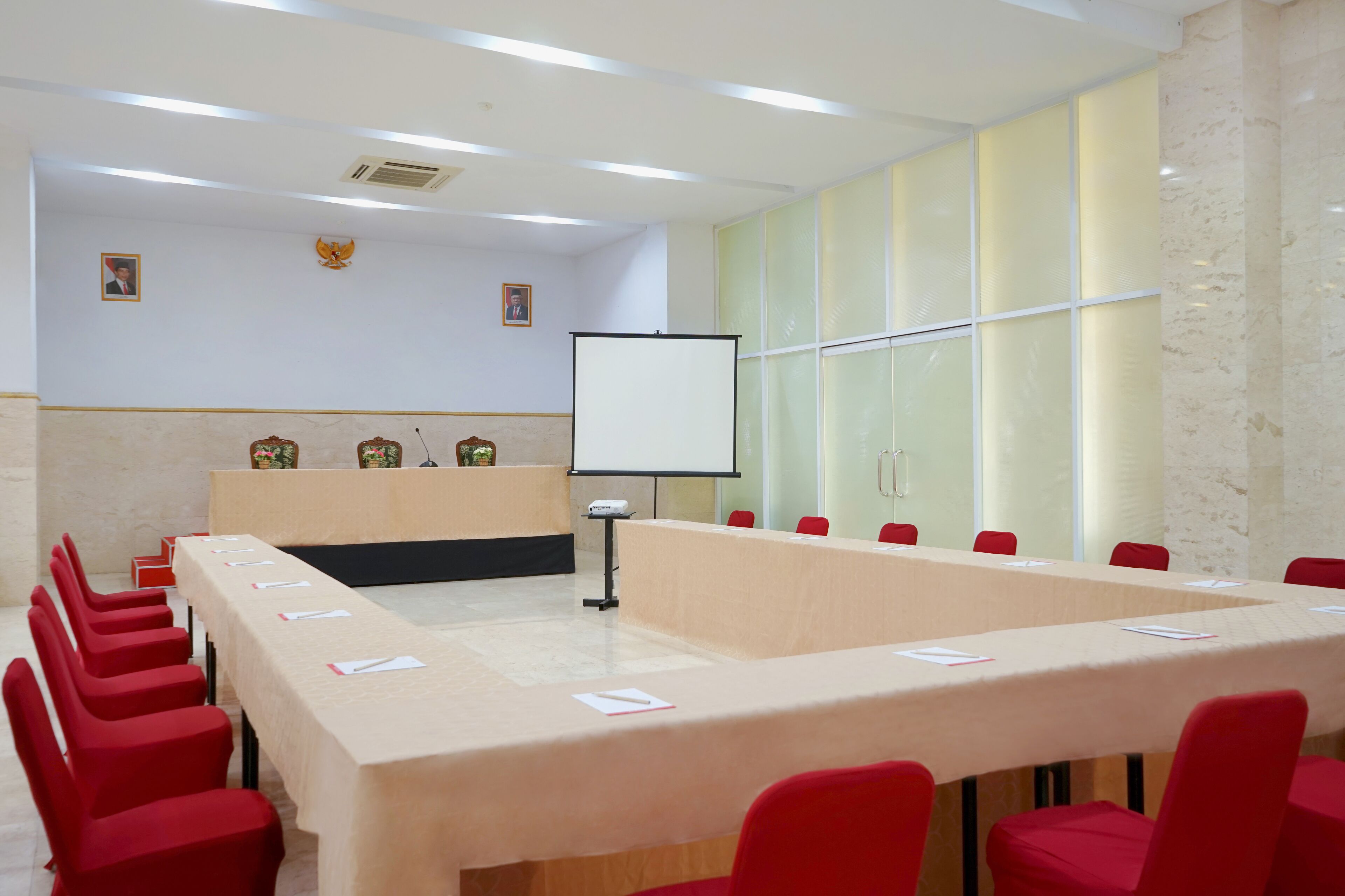 Une salle de conférence avec une longue table, des chaises rouges, un pupitre, un écran de projection et un tableau blanc.