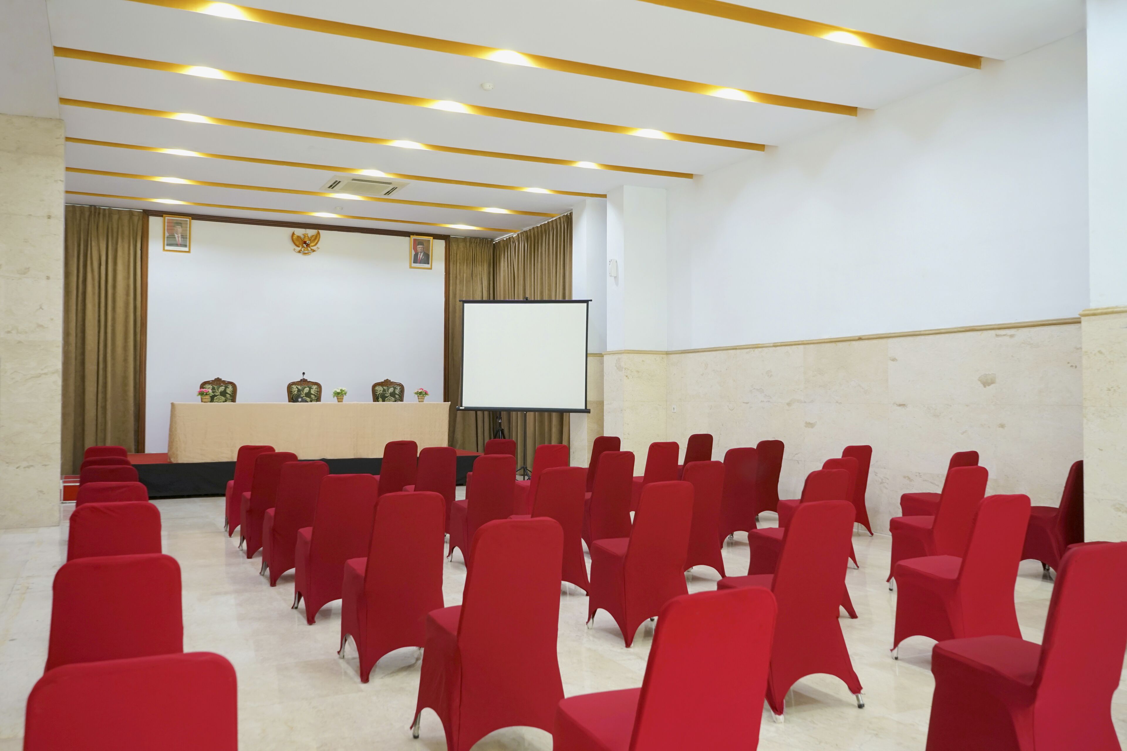 A conference room with rows of red chairs, a podium, a screen, and a stage with a backdrop.