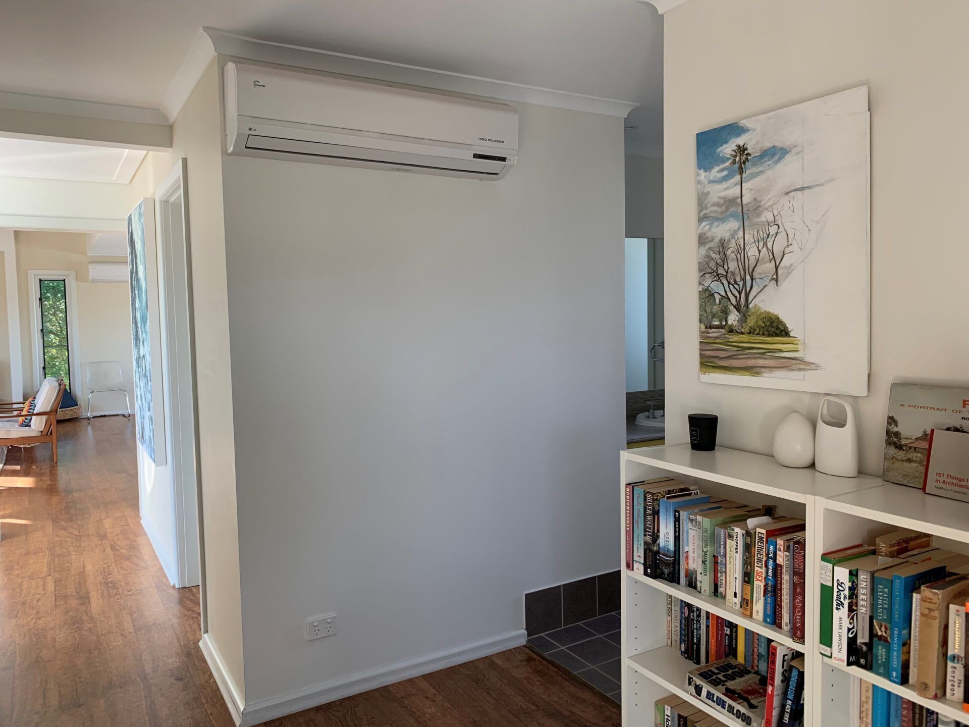 Un salon moderne avec un mur blanc, une bibliothèque remplie de livres, un tableau accroché et un climatiseur fixé au plafond.