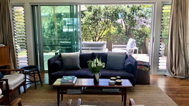 A living room with a dark blue sofa, a coffee table with a vase of flowers, and a view of a garden through large glass doors. - Blauhaus - Relax all your blues away... (Robe)