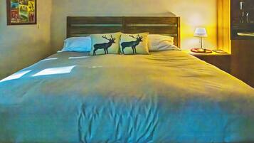 A bed with a headboard, two pillows with deer silhouettes, a bedside table with a lamp, and a wall with a framed picture.