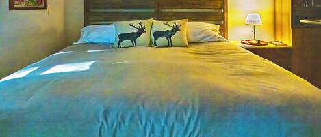 Cama com cabeceira, dois travesseiros com silhuetas de veados, mesa de cabeceira com abajur e parede com uma moldura.