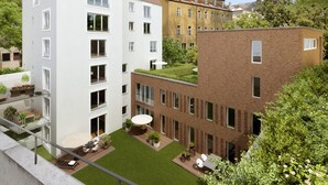 Un moderno complejo de apartamentos con varios edificios, incluyendo una construcción blanca con techo inclinado y otra de ladrillo con techo plano. Hay zonas de estar exteriores con mesas y sillas, y un pequeño jardín con césped y plantas en macetas.