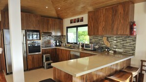 A modern kitchen with wooden cabinets, stainless steel appliances, a large island with stools, and a view of greenery outside.